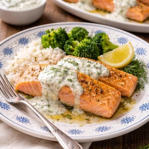 Op de afbeelding: Een bord met gekookte zalmfilets met een romige dillesaus, geserveerd met rijst, broccoli en een citroenpartje. Het bord heeft een blauw en wit bloemmotief. Er ligt een vork op het bord. Op de achtergrond staat een ander bord met zalm.