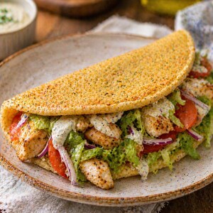 May include: A close-up of a chicken wrap on a speckled plate. The wrap is filled with chicken, lettuce, tomato, and red onion, drizzled with a creamy sauce. The wrap's exterior is golden brown and textured.