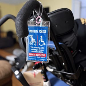 Puede incluir: Una etiqueta rectangular de plástico azul y blanco colgada del asa de un dispositivo de movilidad. La etiqueta muestra el texto MOBILITY ACCESS, un icono de silla de ruedas y el mensaje: This mobility aid is used by a person with a disability.