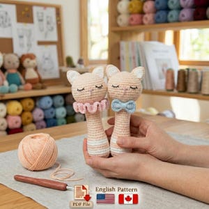 May include: Two handmade crochet cat rattles in peach and white, one with a pink ruffled collar and the other with a blue bow tie. The rattles are held in a person's hands. A ball of peach yarn and a crochet hook are in the foreground.