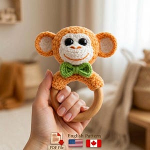 May include: Crocheted monkey rattle in orange, white, and green. The rattle features a wooden ring and a bow tie. The text "English Pattern" is visible, along with a PDF file icon and American and Canadian flags.