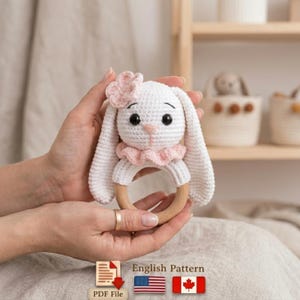 Pode incluir: Um chocalho de bebê de coelho de crochê branco com uma flor e gola rosa. O chocalho tem um anel de madeira e é segurado em duas mãos. A imagem também inclui um ícone de arquivo PDF e o texto "English Pattern".