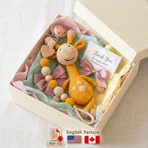 Könnte beinhalten: Ein gehäkeltes Giraffen-Babyspielzeug und Schnullerclip-Set in einer Geschenkbox. Die Giraffe ist gelb mit braunen Flecken. Der Schnullerclip hat Holzperlen und einen herzförmigen Anhänger. Die Box enthält Seidenpapier und eine Dankeskarte.