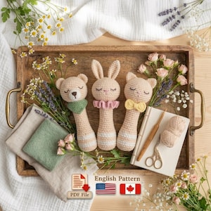 May include: Three handmade crochet baby rattles in the shape of a bear, bunny, and cat, resting on a wooden tray with flowers. The bear has a green scarf, the bunny a pink ruffle, and the cat a yellow bow tie. A PDF file icon and English pattern text are visible.