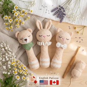 Può includere: Tre sonagli per bambini fatti a mano all'uncinetto a forma di orso, coniglio e gatto. I sonagli sono beige con strisce bianche e hanno tratti del viso ricamati. L'orso ha una sciarpa verde, il coniglio un colletto a balze e il gatto un papillon. L'immagine include il testo "English Pattern".