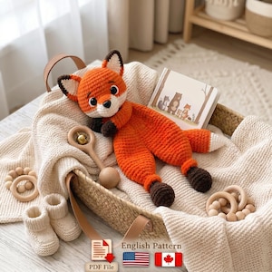May include: A crocheted orange and white fox plush toy with dark brown paws and ears, accompanied by a wooden rattle, two wooden bead rings, and a pair of cream-colored knit booties. A small illustrated book and a PDF file icon are included.