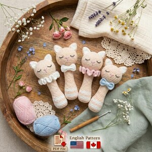 May include: Four handmade crochet cat-shaped baby rattles in soft pastel colors. Each rattle features a cat face with closed eyes, a ruffled collar or bow tie, and striped handle. The rattles are displayed on a wooden tray with yarn, flowers, and a crochet hook.