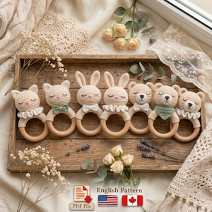 May include: A collection of six handmade crochet baby rattles in the shape of animals, including bears, bunnies, and cats. Each rattle features a wooden ring and is adorned with a bow tie or collar. The rattles are displayed on a wooden tray.