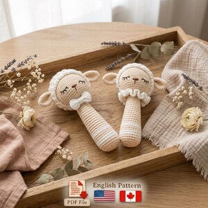 May include: Two handmade crochet sheep rattles in cream and beige, with closed eyes and a bow tie. The rattles are placed on a wooden tray with floral and fabric accents. The image also includes a PDF file icon and the text "English Pattern".
