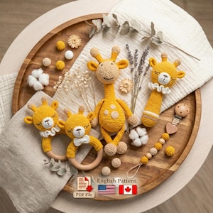 Könnte beinhalten: Eine Sammlung handgefertigter Baby-Spielzeuge aus Häkelarbeit, darunter Giraffen-Rasseln und Beißringe, in Gelb- und Beigetönen. Die Spielzeuge sind auf einem Holztablett mit dekorativen Elementen wie Trockenblumen und Baumwolle angeordnet. Der Text "English Pattern" ist enthalten.