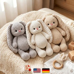 May include: Three plush crochet bunnies in gray, white, and beige. Each stuffed animal features long floppy ears, dark round eyes, and a small pink nose. The toys are crafted from a soft, textured yarn and are positioned side by side.