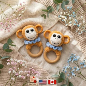 May include: Two handmade crochet monkey baby rattles. Each rattle features a smiling monkey face with orange fur, a white face, black eyes, and a blue ruffled collar. The monkey is attached to a wooden ring. The background includes flowers and a knitted blanket.
