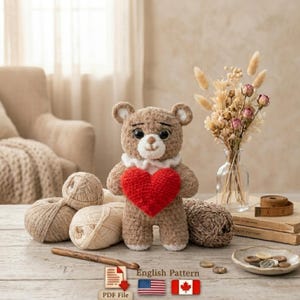 May include: A handmade crochet teddy bear with a red heart, wearing a white collar. The bear is brown with black eyes and a white snout. Several balls of yarn and a crochet hook are in the foreground. The text "English Pattern" is visible.