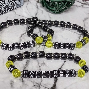 May include: Two black beaded bracelets with yellow smiley face beads. The bracelets spell out "CREATE YOUR HAPPINESS".