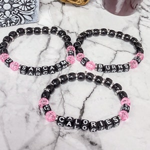 May include: Three black beaded bracelets with pink beads and black letter tiles spelling out "Sarcasm", "Burns", and "Calories".