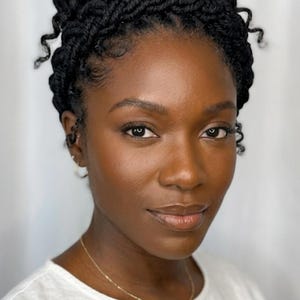 May include: A portrait of a person with dark skin and black braided hair styled in a high bun. The person is wearing a white top and a delicate gold necklace. The background is a plain white.