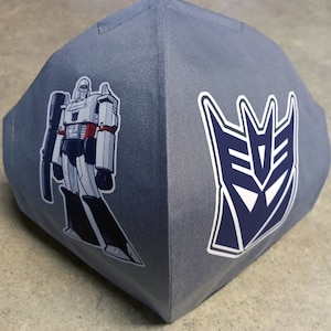 May include: Gray face mask with a cartoon image of the Transformers character Megatron and the Autobot symbol.
