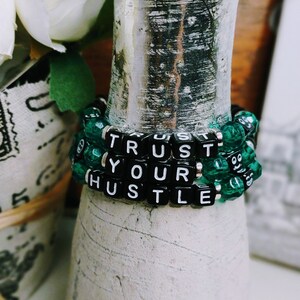 May include: A green and black beaded bracelet with the words "TRUST YOUR HUSTLE" spelled out in black letter beads.