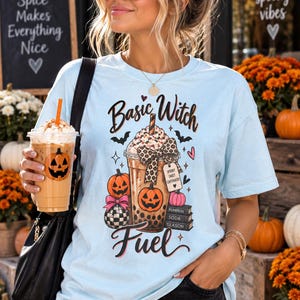 Puede incluir: Una camiseta azul claro con un estampado gráfico de una taza de café con un lazo de leopardo, calabazas y el texto Basic Witch Fuel. El diseño incluye una etiqueta con la inscripción Spooky Sweet Caffeinated y libros con Pumpkin Soda Season.