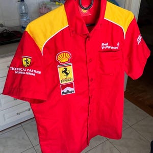 Könnte beinhalten: Rotes kurzärmeliges Hemd mit gelben Schulterpartien. Enthält Ferrari-, Shell V-Power- und Marlboro-Logos. Das Hemd ist zugeknöpft und wird von einem schwarzen Kleiderbügel gehalten. Ein klassisches Motorsport-Thema.