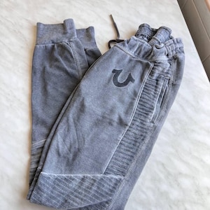 May include: A pair of grey sweatpants featuring a faded wash, an elastic waistband with a drawstring, and ribbed panels on the sides and lower legs. The True Religion horseshoe logo is embroidered in a dark grey thread on the upper front thigh.