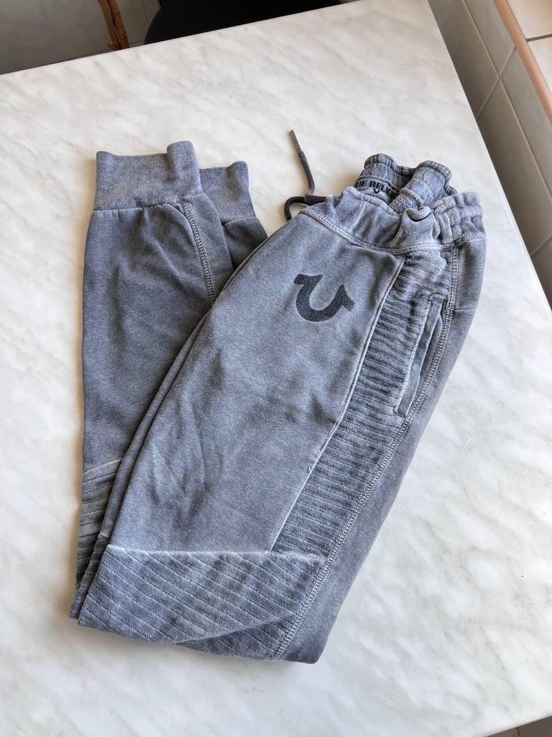 May include: A pair of grey sweatpants featuring a faded wash, an elastic waistband with a drawstring, and ribbed panels on the sides and lower legs. The True Religion horseshoe logo is embroidered in a dark grey thread on the upper front thigh.