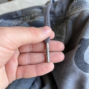 May include: A grey fabric garment featuring a thick grey drawstring with a metallic silver aglet. The cylindrical metal tip is engraved with a small, detailed emblem. The fabric has a textured, slightly faded appearance with visible stitching.