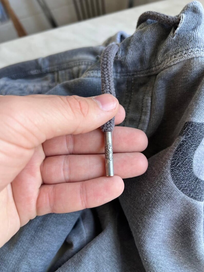 May include: A grey fabric garment featuring a thick grey drawstring with a metallic silver aglet. The cylindrical metal tip is engraved with a small, detailed emblem. The fabric has a textured, slightly faded appearance with visible stitching.