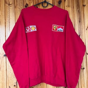 Könnte beinhalten: Ein rotes Sweatshirt mit Aufnähern von Vodafone, Shell und Marlboro. Das Sweatshirt mit Rundhalsausschnitt hat lange Ärmel und hängt an einem schwarzen Kleiderbügel. Die Aufnäher befinden sich auf der oberen Brust. Der Hintergrund ist aus Holz.