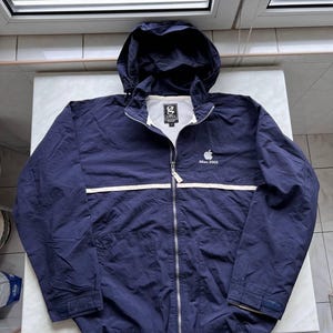 Puede incluir: Chaqueta azul marino con capucha y una franja blanca horizontal en el pecho. La chaqueta tiene cierre de cremallera, un pequeño logotipo de Apple y el texto "iMac 2005". Tiene forro blanco y un pequeño logotipo en el pecho.