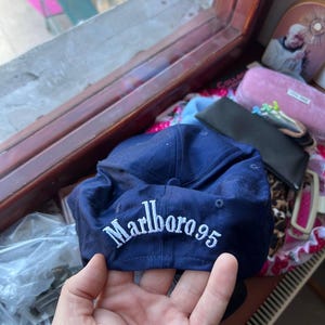 Op de afbeelding: Een marineblauwe baseballpet met de wit geborduurde tekst Marlboro 95 op het voorpaneel. De pet is gemaakt van een stevige stof met een gestructureerde kroon en een knoop aan de bovenzijde, voorzien van duidelijke witte stikseldetails.