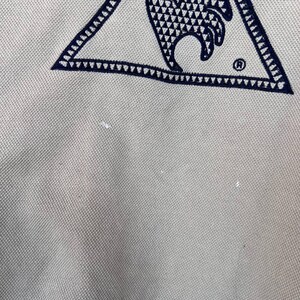 Puede incluir: Primer plano de una tela beige claro con un logotipo triangular azul marino. El logotipo presenta un dise&ntilde;o de gallo con un patr&oacute;n geom&eacute;trico y un s&iacute;mbolo de marca registrada.