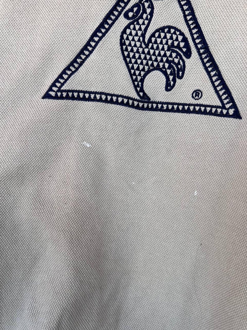 Puede incluir: Primer plano de una tela beige claro con un logotipo triangular azul marino. El logotipo presenta un dise&ntilde;o de gallo con un patr&oacute;n geom&eacute;trico y un s&iacute;mbolo de marca registrada.