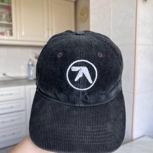 Op de afbeelding: Een zwarte corduroy baseballpet met een wit geborduurd Aphex Twin-logo in het midden van het voorpaneel. De pet heeft een gestructureerde kroon, een gebogen klep en een zichtbare verticale corduroy textuur over de gehele stof.