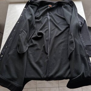 May include: Black jacket with a zip-up front, snap closures, and two front pockets. The jacket is open, revealing the interior lining and a small orange tag near the collar. The jacket appears to be made of a soft, dark fabric.