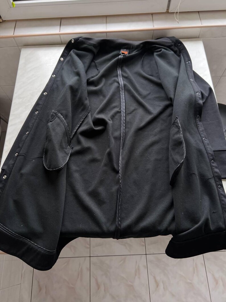 May include: Black jacket with a zip-up front, snap closures, and two front pockets. The jacket is open, revealing the interior lining and a small orange tag near the collar. The jacket appears to be made of a soft, dark fabric.