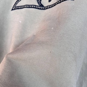 Puede incluir: Primer plano de una tela verde claro con un emblema azul marino en la esquina superior izquierda. El emblema es un tri&aacute;ngulo con un dise&ntilde;o de ola en el interior. La tela tiene una apariencia texturizada.