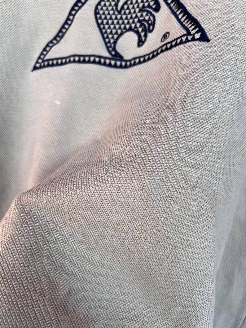 Puede incluir: Primer plano de una tela verde claro con un emblema azul marino en la esquina superior izquierda. El emblema es un tri&aacute;ngulo con un dise&ntilde;o de ola en el interior. La tela tiene una apariencia texturizada.