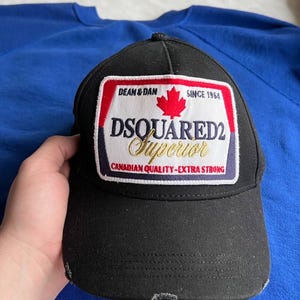Może przedstawiać: Czarna czapka z daszkiem z postrzępionym brzegiem. Czapka posiada prostokątną naszywkę z napisem "DSQUARED2 Superior" i czerwoną liściem klonu. Naszywka zawiera również tekst "DEAN & DAN SINCE 1964" i "CANADIAN QUALITY-EXTRA STRONG."