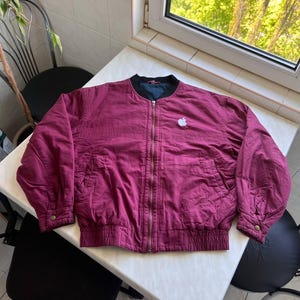 May include: A maroon bomber jacket featuring a full-length front zipper, a black ribbed collar, and a white Apple logo embroidered on the left chest. The jacket includes long sleeves with buttoned cuffs and a ribbed hem, with a dark interior lining.