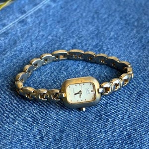 May include: A gold-coloured wristwatch with a rectangular face and a linked bracelet. The watch face is white with black hands and the brand name "Q&Q" is visible. The watch is displayed on a blue denim surface.