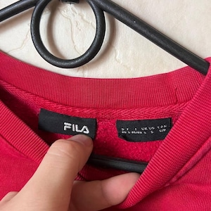 May include: A close-up of a red crew neck sweatshirt with the brand name "FILA" in white on a black tag. The sweatshirt is hanging on a black hanger. The tag also includes size information.