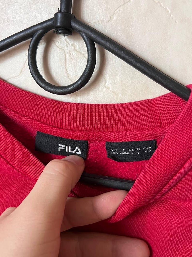 May include: A close-up of a red crew neck sweatshirt with the brand name "FILA" in white on a black tag. The sweatshirt is hanging on a black hanger. The tag also includes size information.