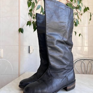 May include: A pair of black leather knee-high boots featuring a rounded toe, a low stacked heel, and a slouchy shaft design. The boots have visible stitching along the foot and a smooth, matte finish on the leather surface, extending to the calf.