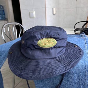 Puede incluir: Un sombrero de pescador azul marino con ala ancha y un parche ovalado verde en la parte frontal con el texto LAND ROVER bordado en amarillo. El sombrero incluye ojales metálicos en los laterales y una correa para la barbilla.