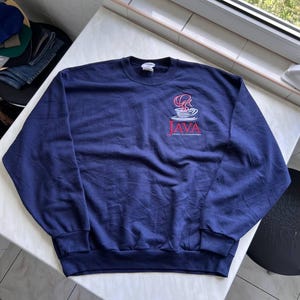 May include: A navy blue crewneck sweatshirt featuring a graphic on the upper left chest. The graphic depicts a steaming cup of coffee in white and red, with the word "JAVA" printed in red capital letters below it, followed by smaller text.