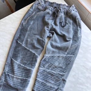 May include: Gray sweatpants featuring a faded, washed-out finish, an elastic waistband with a drawstring, and ribbed paneling on the thighs and knees. A dark horseshoe logo is embroidered on the upper left thigh area of the garment.
