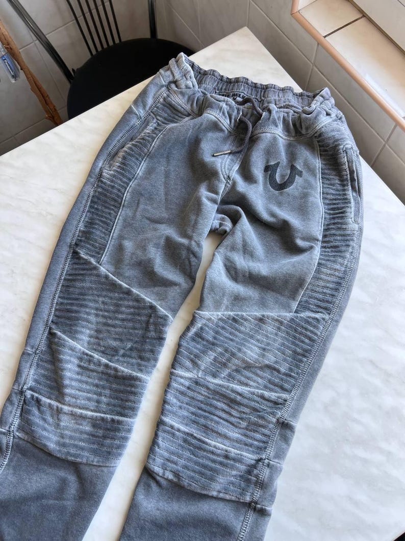 May include: Gray sweatpants featuring a faded, washed-out finish, an elastic waistband with a drawstring, and ribbed paneling on the thighs and knees. A dark horseshoe logo is embroidered on the upper left thigh area of the garment.