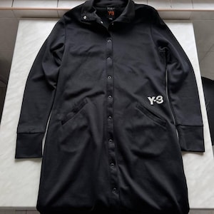 May include: Black long-sleeve button-down shirt with a collar and two front pockets. The shirt features the white "Y-3" logo on the front and a small "Y-3" logo on the collar. The cuffs are ribbed.