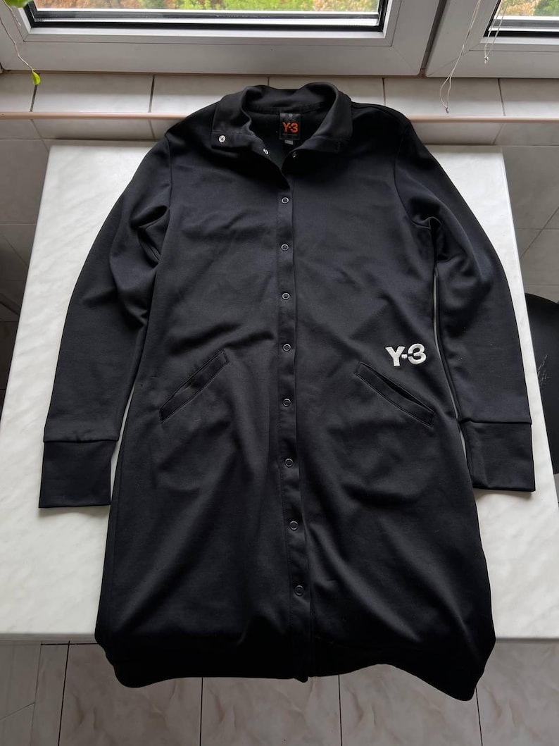 May include: Black long-sleeve button-down shirt with a collar and two front pockets. The shirt features the white "Y-3" logo on the front and a small "Y-3" logo on the collar. The cuffs are ribbed.